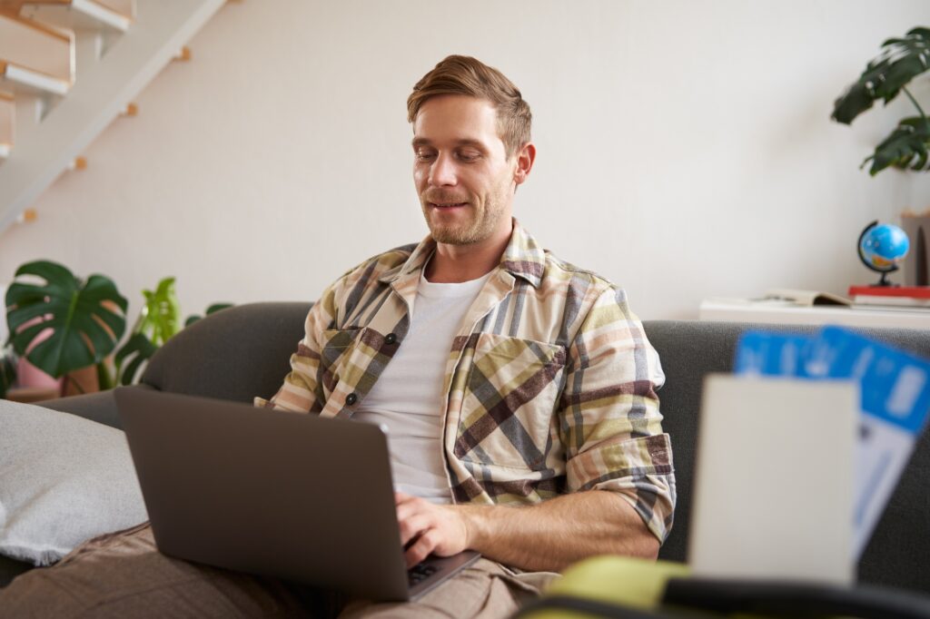 Portrait of handsome young man, has two tickets and passport, goes online on laptop, booking hotel